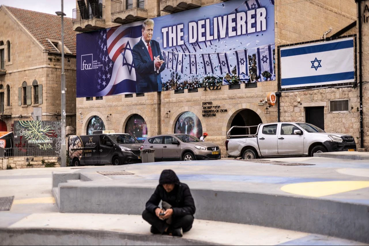 A man sits in front of a banner depicting US President Donald Trump with the slogan “The Deliverer” in Jerusalem on April 20, 2026. (AFP Photo)