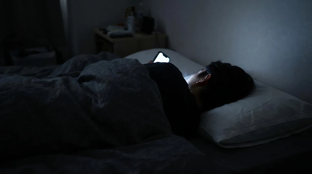 A person uses a smartphone while lying in bed at night, illuminated by the screen. (Adobe Stock Photo)