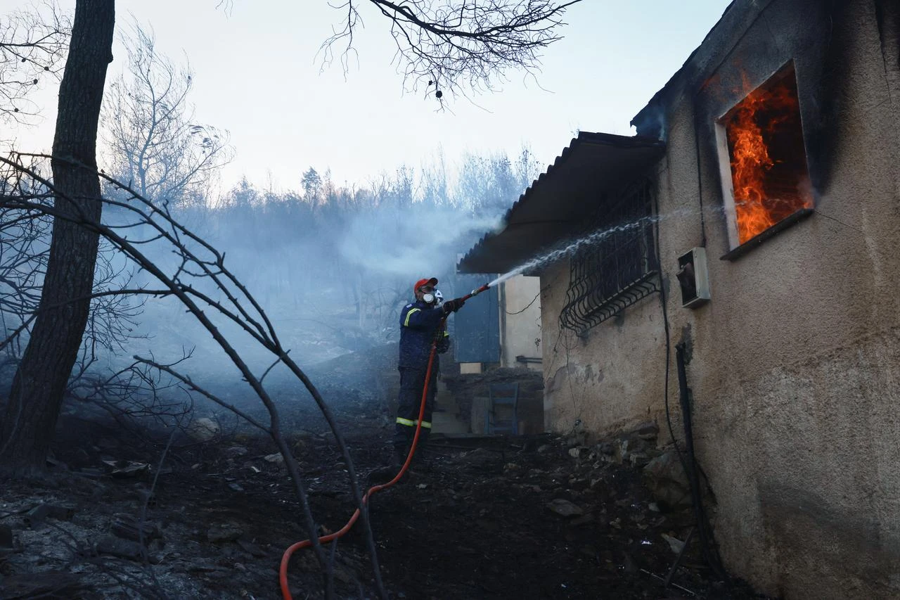 A firefighter sprays water on a burning house as crews battle a wildfire in Peristeria on Salamina Island, near Athens, Greece, Aug. 16, 2026. (AA Photo)