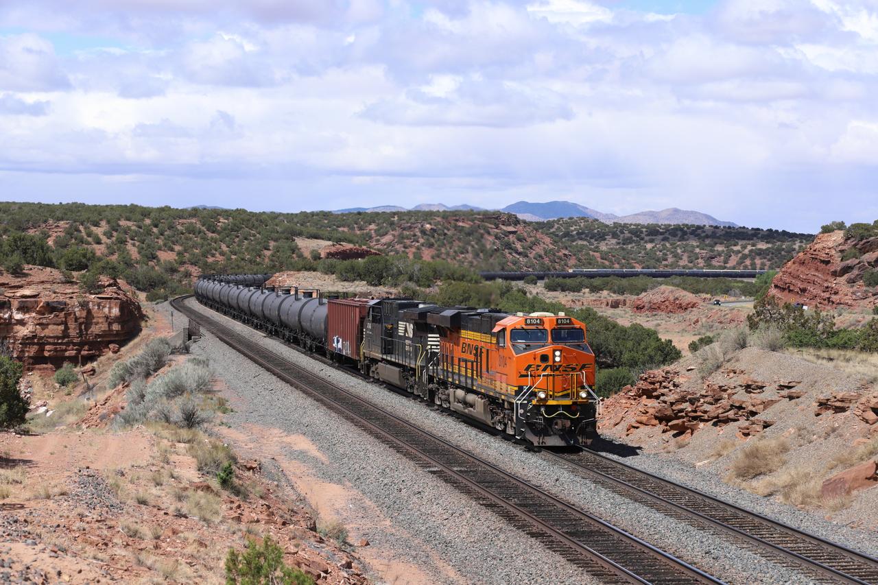 An orange and black BNSF Railway diesel-electric locomotive leads a mixed freight train, including tank cars and boxcars, through a rugged desert canyon in the American Southwest.