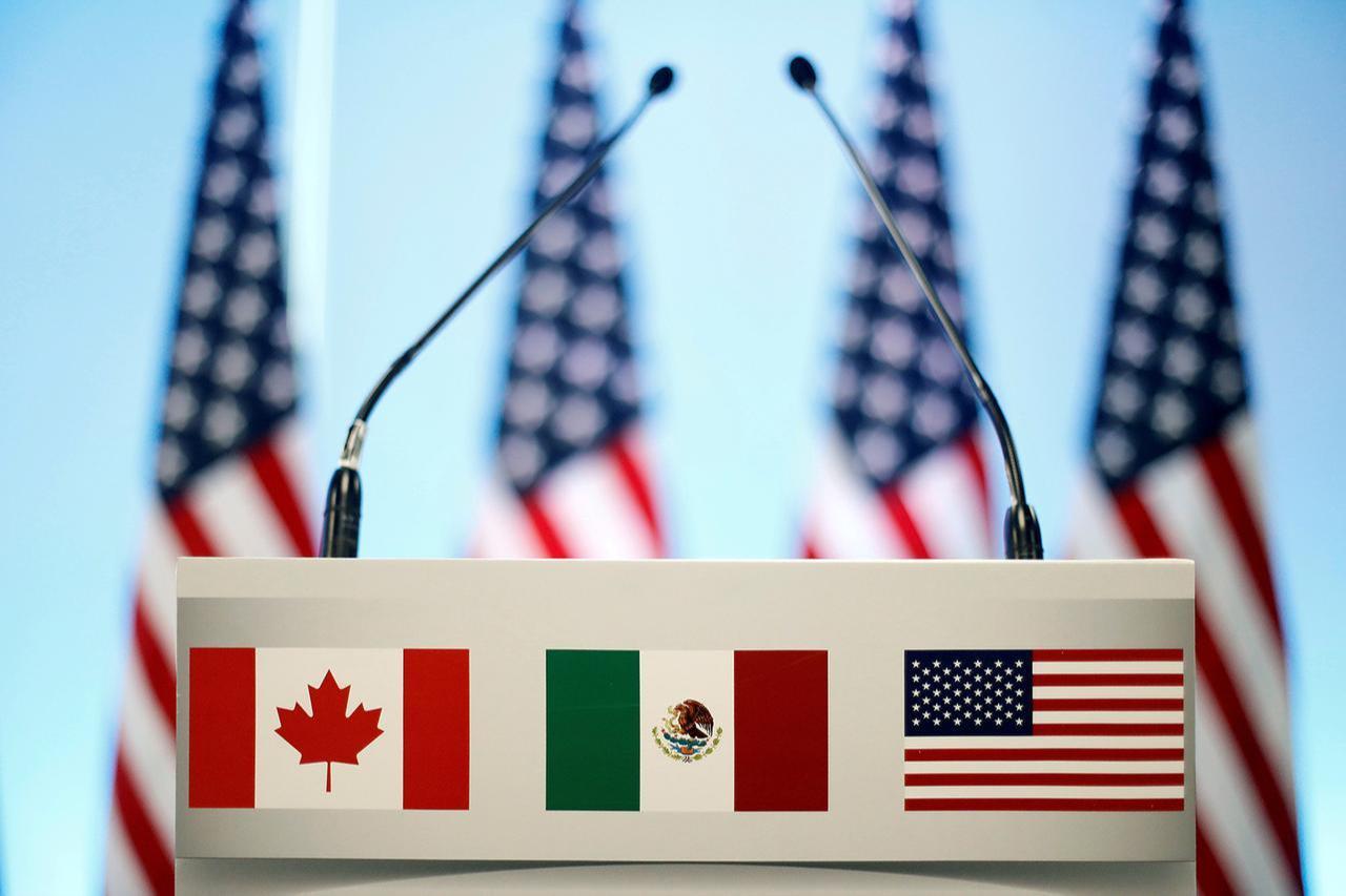 Four flags of the United States frame a white podium displaying the flags of Canada, Mexico, and the United States ahead of North American trade talks.