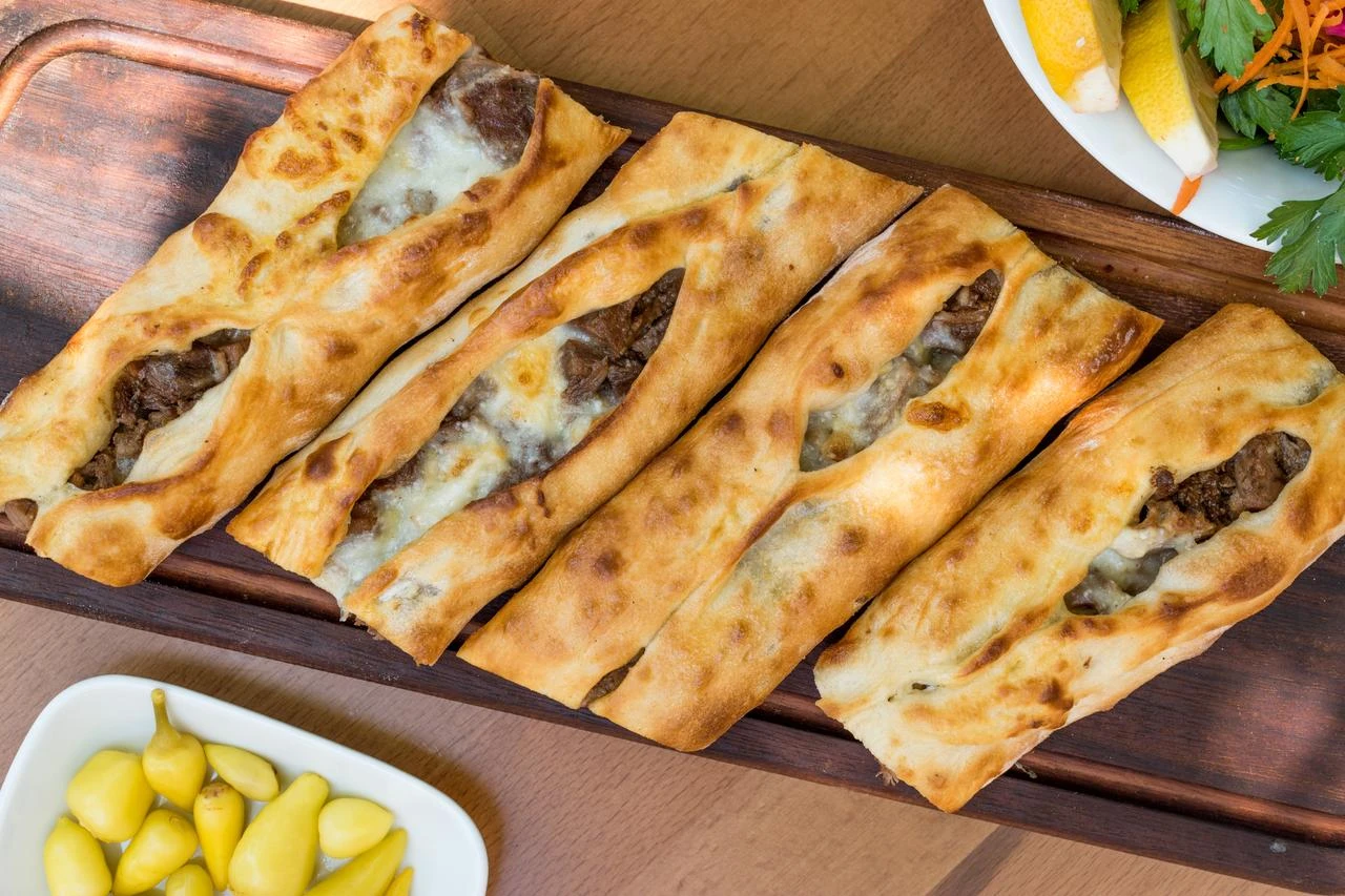 Long, folded Bafra pide filled with diced meat and melted cheese is served on a wooden board with pickled peppers. (Adobe Stock Photo)