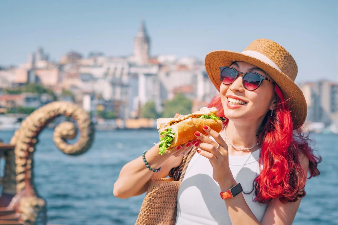 A tourist enjoys fish sandwich (balik ekmek) by the Bosphorus, with Istanbul’s Galata Tower rising in the background. (Adobe Stock Photo)