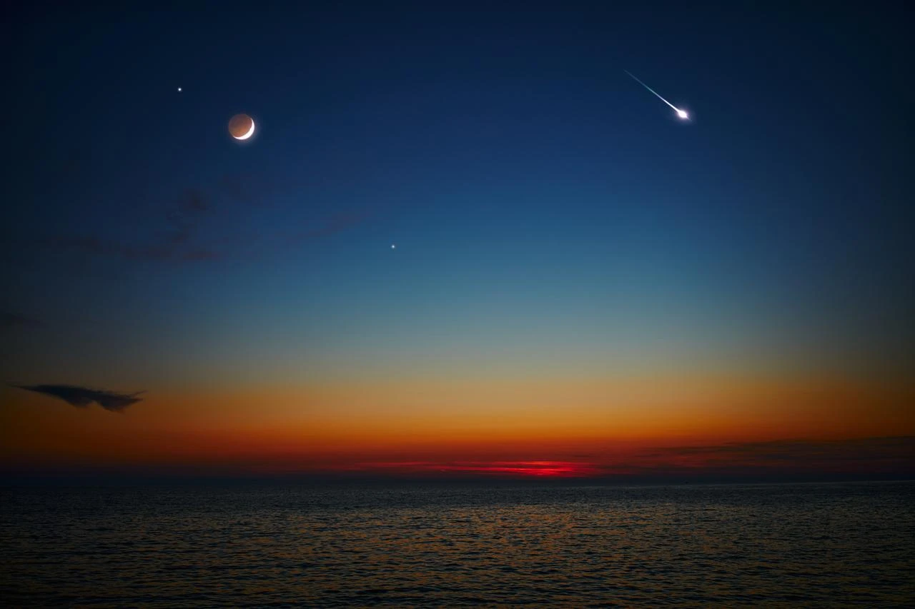A meteor streaks across the sky near a crescent moon above the sea at twilight. (Adobe Stock Photo)
