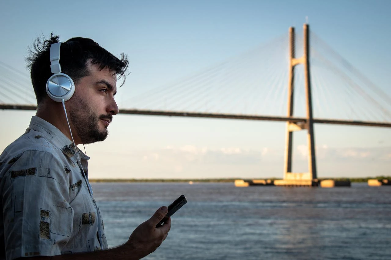 A man listens to audio through headphones while using a smartphone beside a waterfront and cable-stayed bridge. (Adobe Stock Photo)