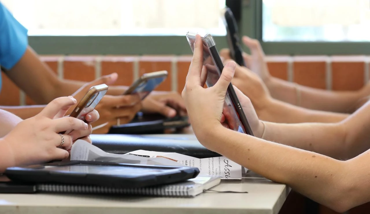 A table of high school students all holding multiple digital devices such as phones and tablets during a class lesson. (Photo via Adobe Stock)