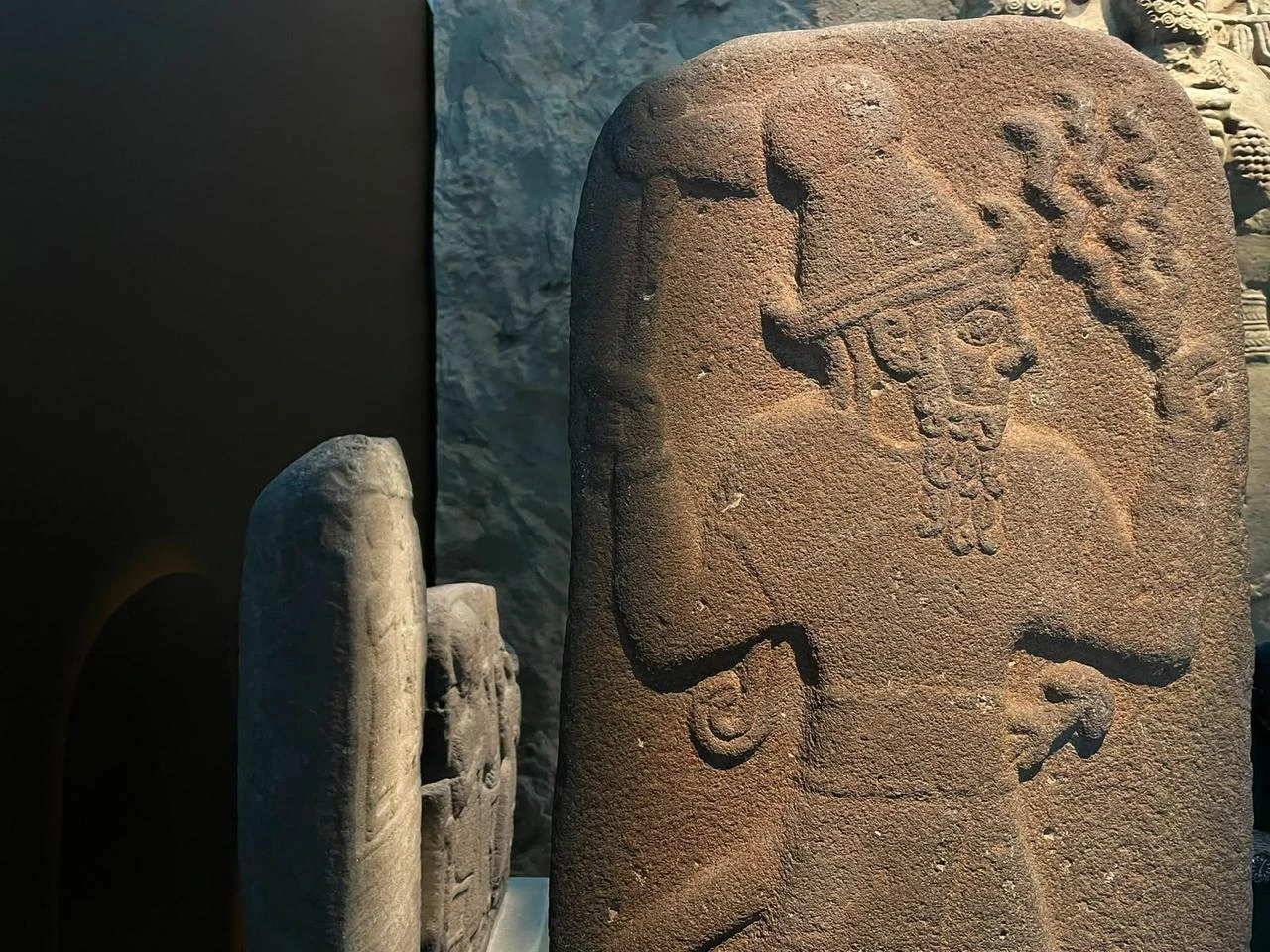 A carved stone relief depicting an ancient figure wearing an elaborate headdress is displayed under focused gallery lighting. (Photo by Koray Erdogan/Türkiye Today)