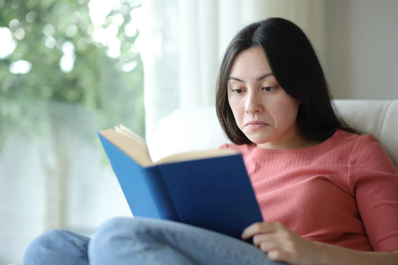 A woman reads a book while seated near a window in this undated stock photo. (Adobe Stock Photo)