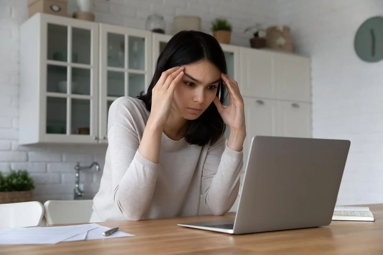 A concerned woman holds her temples while working on a laptop at home. (Adobe Stock Photo)