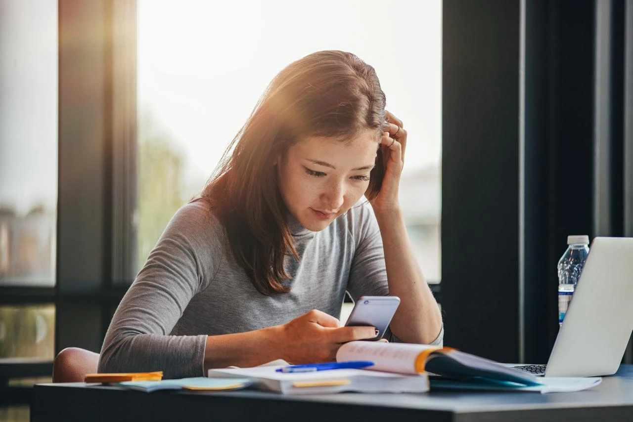 A student checks her smartphone while studying with books and a laptop. (Adobe Stock Photo)