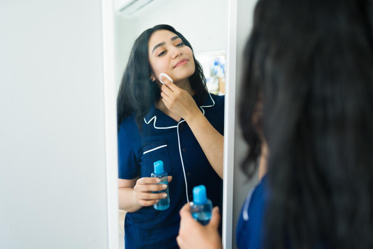 A woman uses a cotton pad and skincare product while looking in a mirror, illustrating personal care and beauty routines. (Adobe Stock Photo)