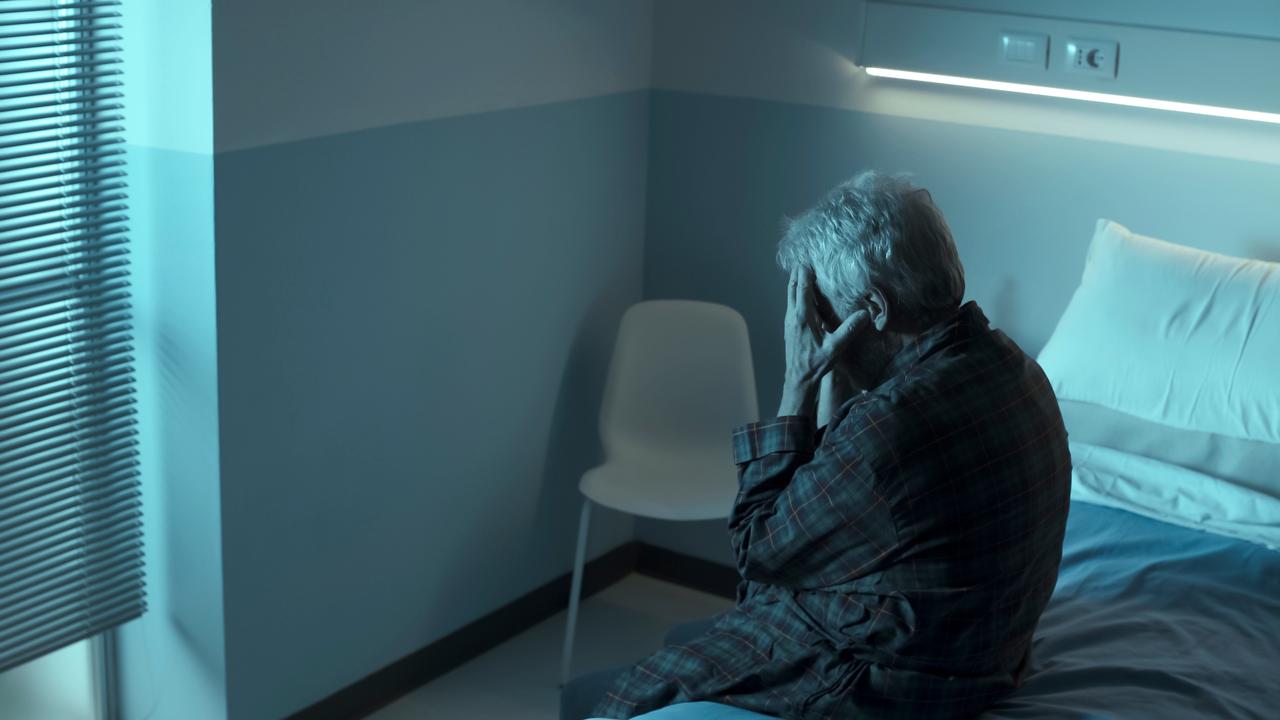 An older man sits on the edge of a bed with his head in his hands. (Adobe Stock Photo)