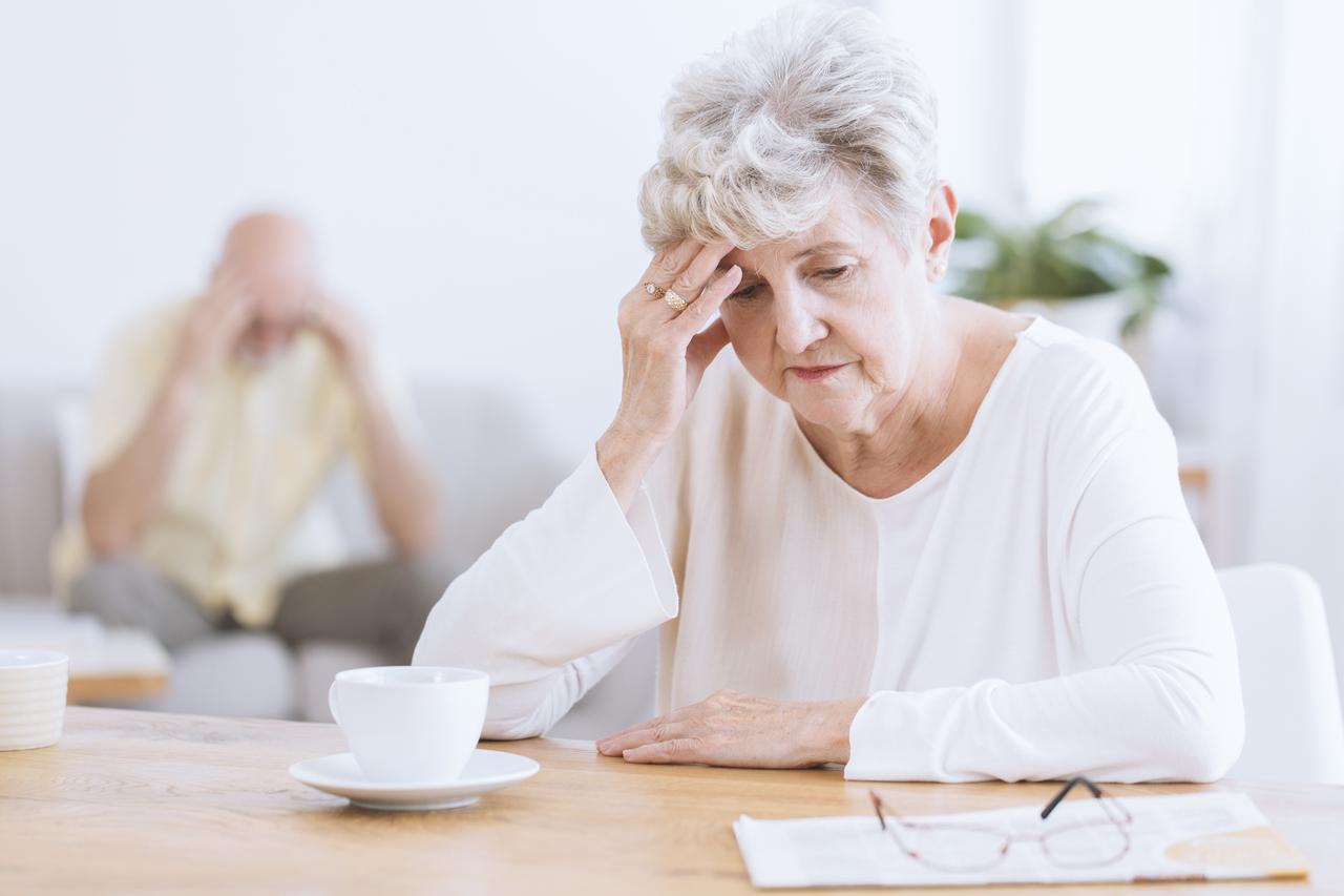 An older woman sits at a table with her hand on her forehead. (Adobe Stock Photo)