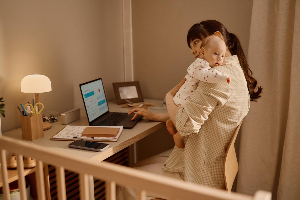 A mother works on a laptop while holding her baby at home. (Adobe Stock Photo)