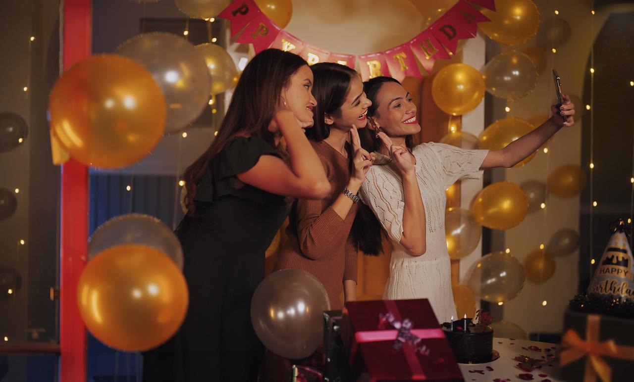 Three women take a selfie during a birthday celebration decorated with balloons and party lights. (Adobe Stock Photo)