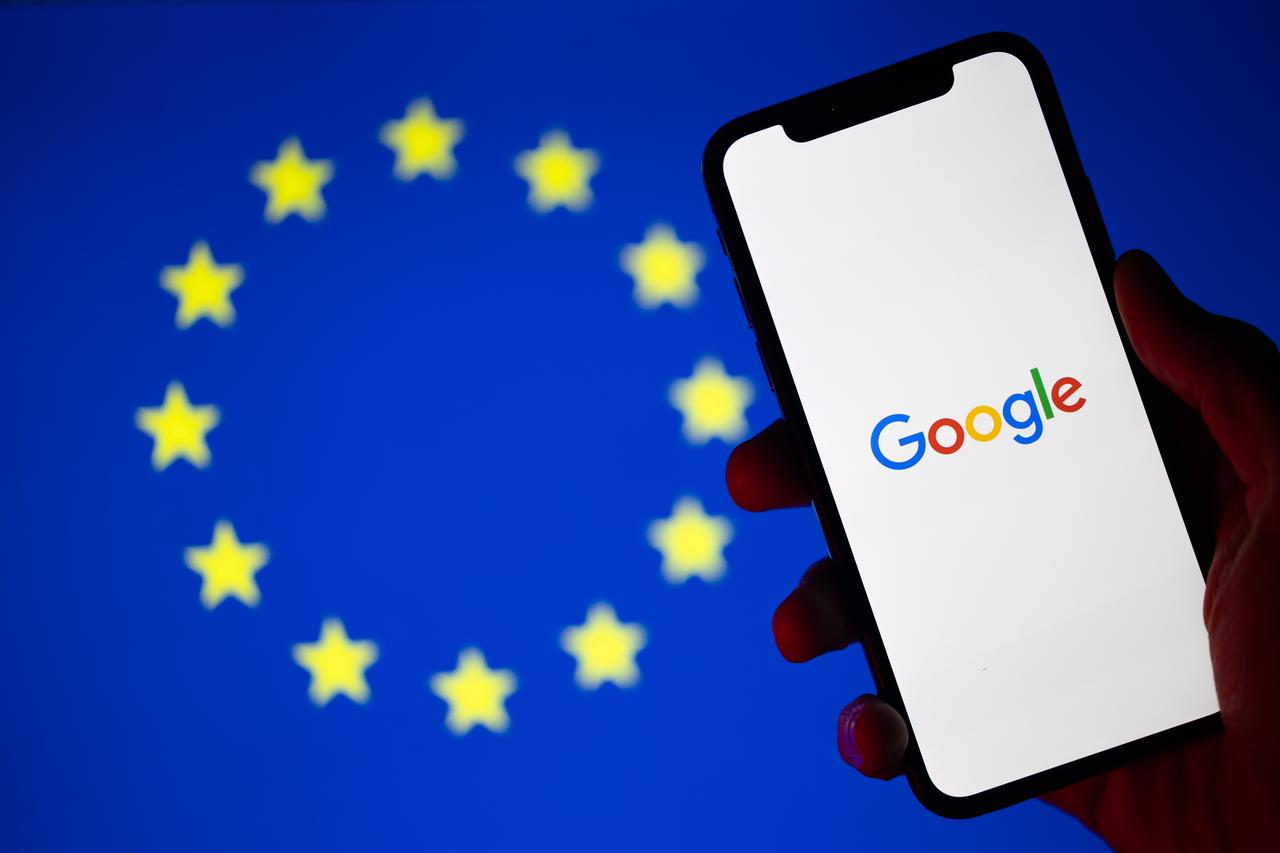 In silhouette, a hand holds a mobile phone with the Google logo displayed. Behind it, a blurred backdrop illustrates the flag of the European Union. (Photo via Adobe Stock)