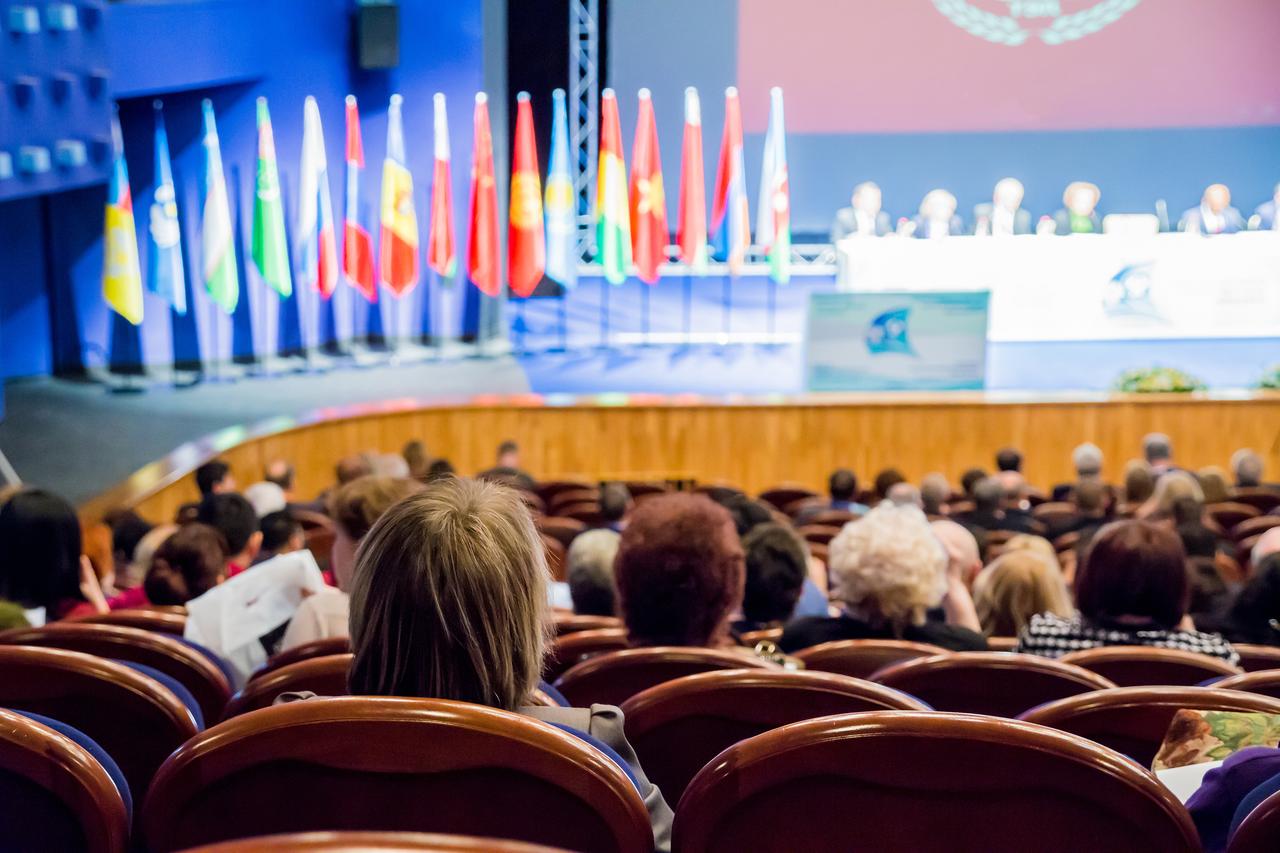 Attendees sit in an auditorium during an international conference with national flags displayed near the stage. (Adobe Stock Photo)