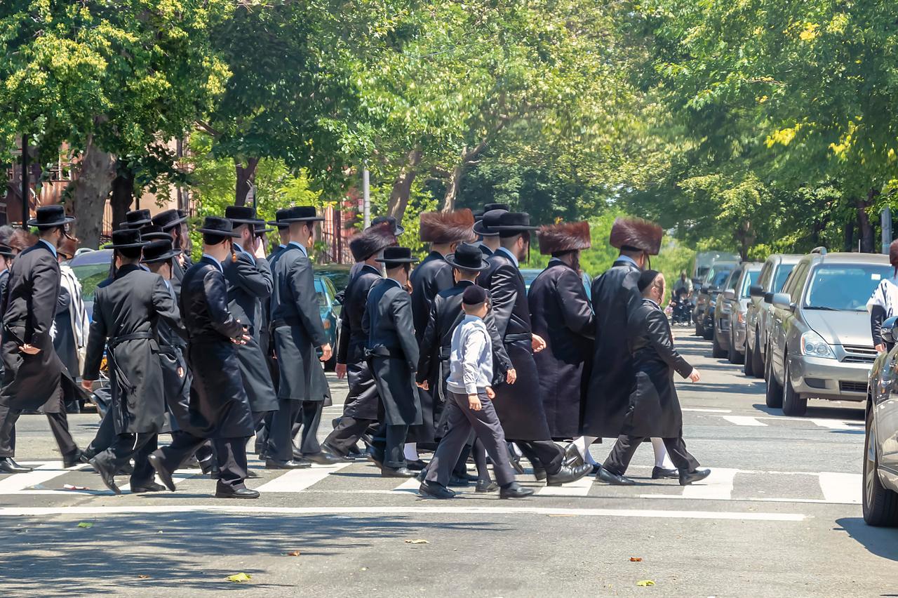 Men and a boy dressed in traditional black attire cross a tree-lined street in Brooklyn, New York. (Adobe Stock Photo)