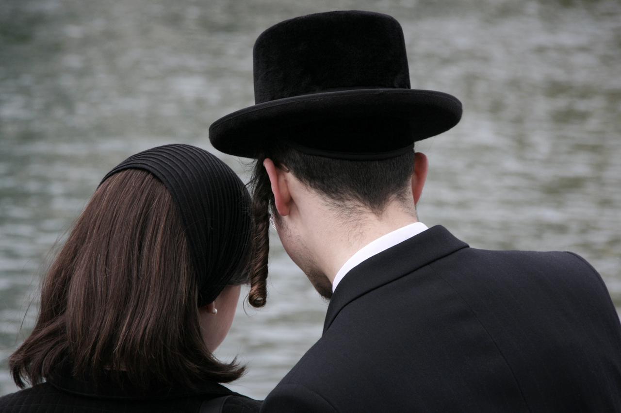 A man and woman in traditional black attire stand together near the waterfront. (Adobe Stock Photo)