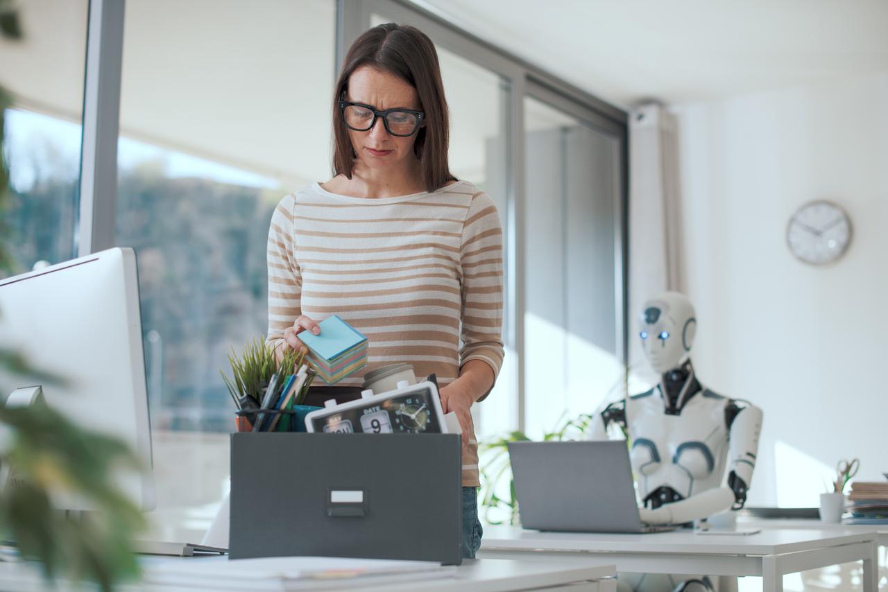 A woman packs belongings into a box at an office desk while a humanoid robot works nearby, illustrating concerns over automation and AI-driven job displacement. (Adobe Stock Photo)